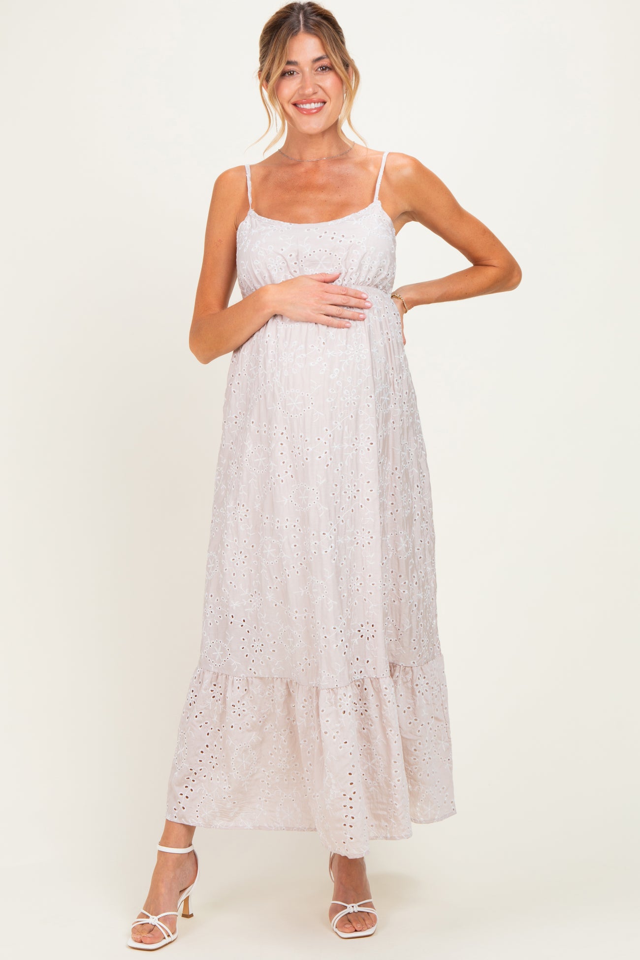 woman wearing a light pink maternity dress with eyelet details, holding her baby bump, featuring a tiered skirt and spaghetti straps