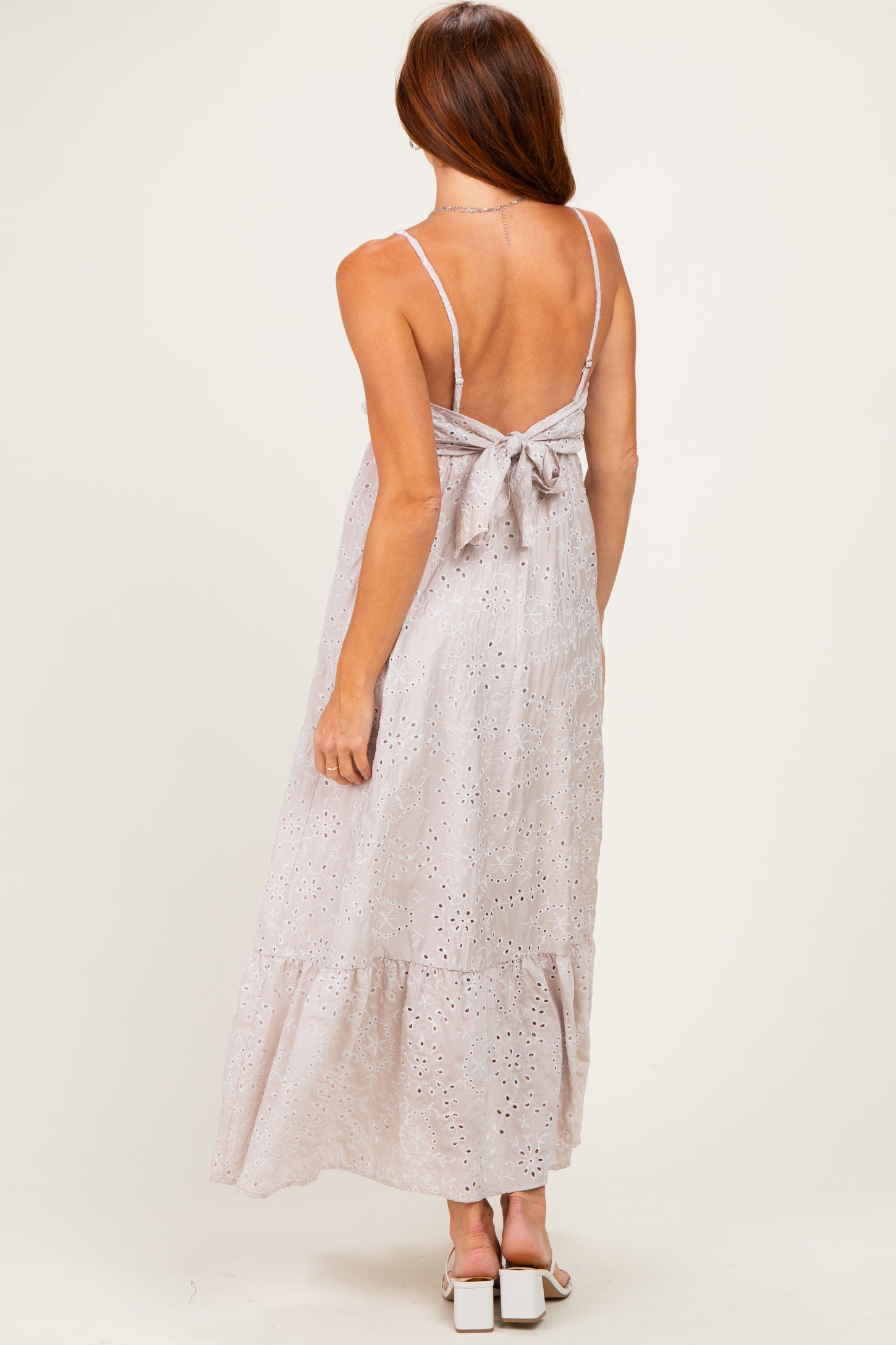 woman wearing a light pink backless maxi dress with a bow detail and eyelet design, posed from the back