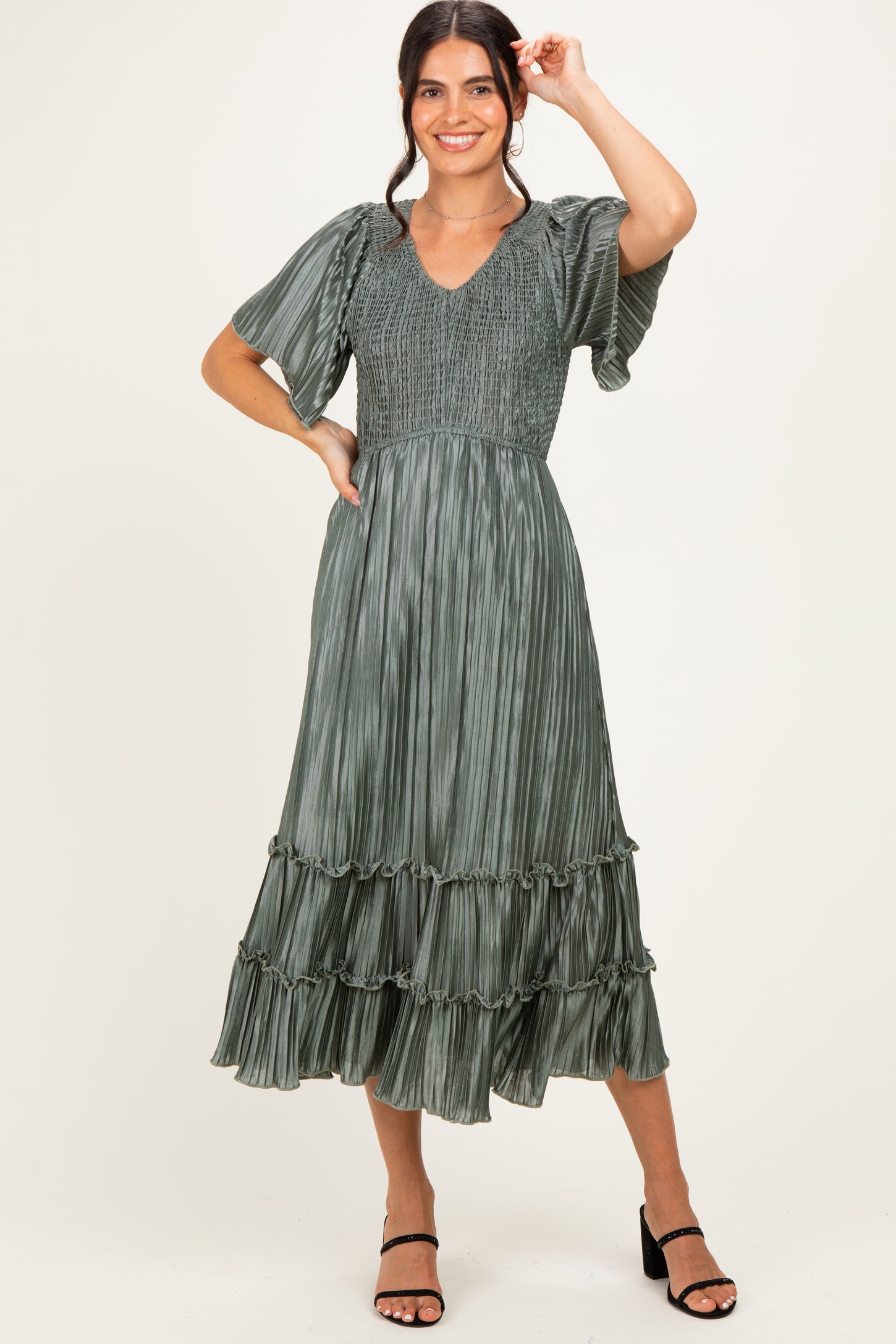 woman wearing a green pleated midi dress with short sleeves and a ruffled hem, smiling and posing with one hand raised