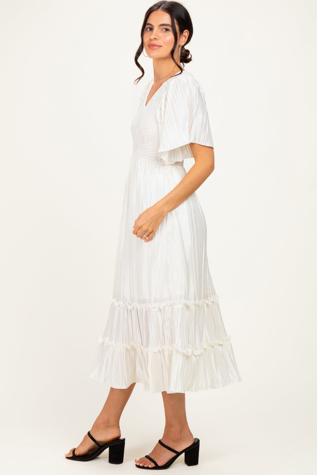 woman wearing a white pleated midi dress with flutter sleeves and ruffled hem, standing sideways with a smile