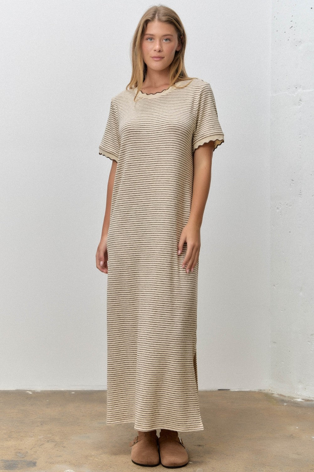 woman wearing a beige striped maxi dress with scalloped edges and side slits, standing on a concrete floor