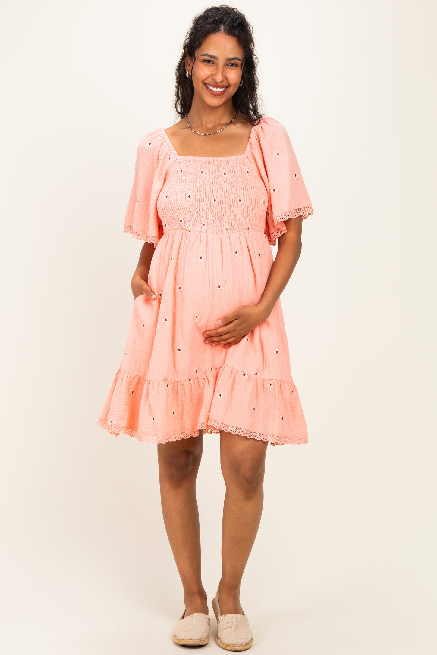 woman wearing a coral maternity dress with short puff sleeves, smocked bodice, and tiered ruffle skirt, holding her baby bump