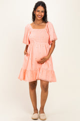 woman wearing a coral maternity dress with short puff sleeves, smocked bodice, and tiered ruffle skirt, holding her baby bump