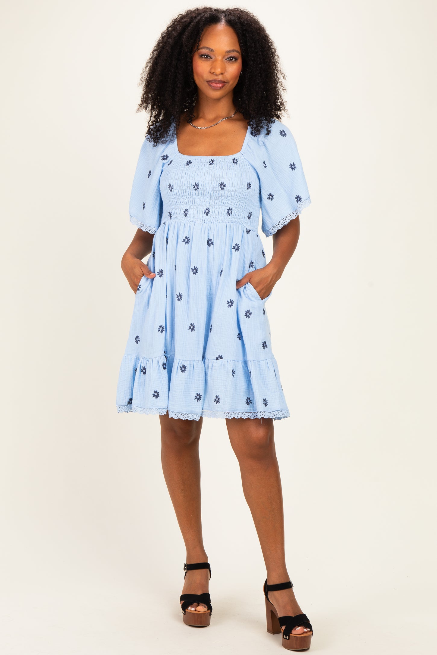 woman wearing a light blue short sleeve dress with floral embroidery and pockets, standing with one hand on her hip