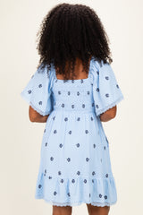 woman wearing a light blue short sleeve dress with floral designs and ruffled hem, seen from the back
