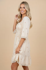 woman wearing a light beige embroidered dress with three-quarter sleeves and lace trim, smiling at the camera