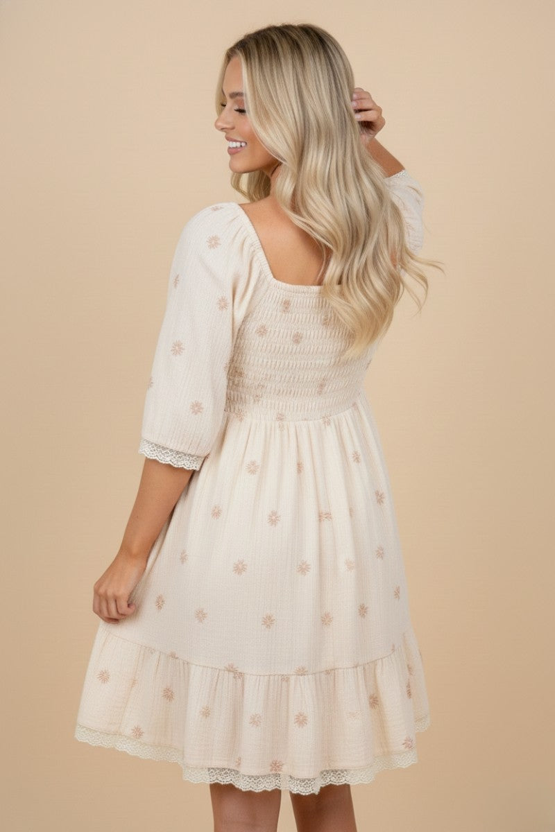 woman wearing a cream-colored smocked dress with floral details and a ruffled hem, viewed from the back