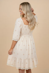 woman wearing a cream-colored smocked dress with floral details and a ruffled hem, viewed from the back