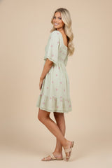 woman wearing a light green short sleeve dress with floral embroidery and a tiered ruffle hem, standing sideways