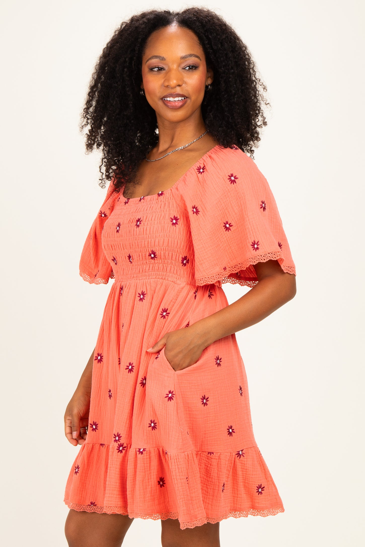 woman wearing a coral short puff sleeve dress with floral embroidery and a tiered ruffle hem, posing with one hand in the pocket