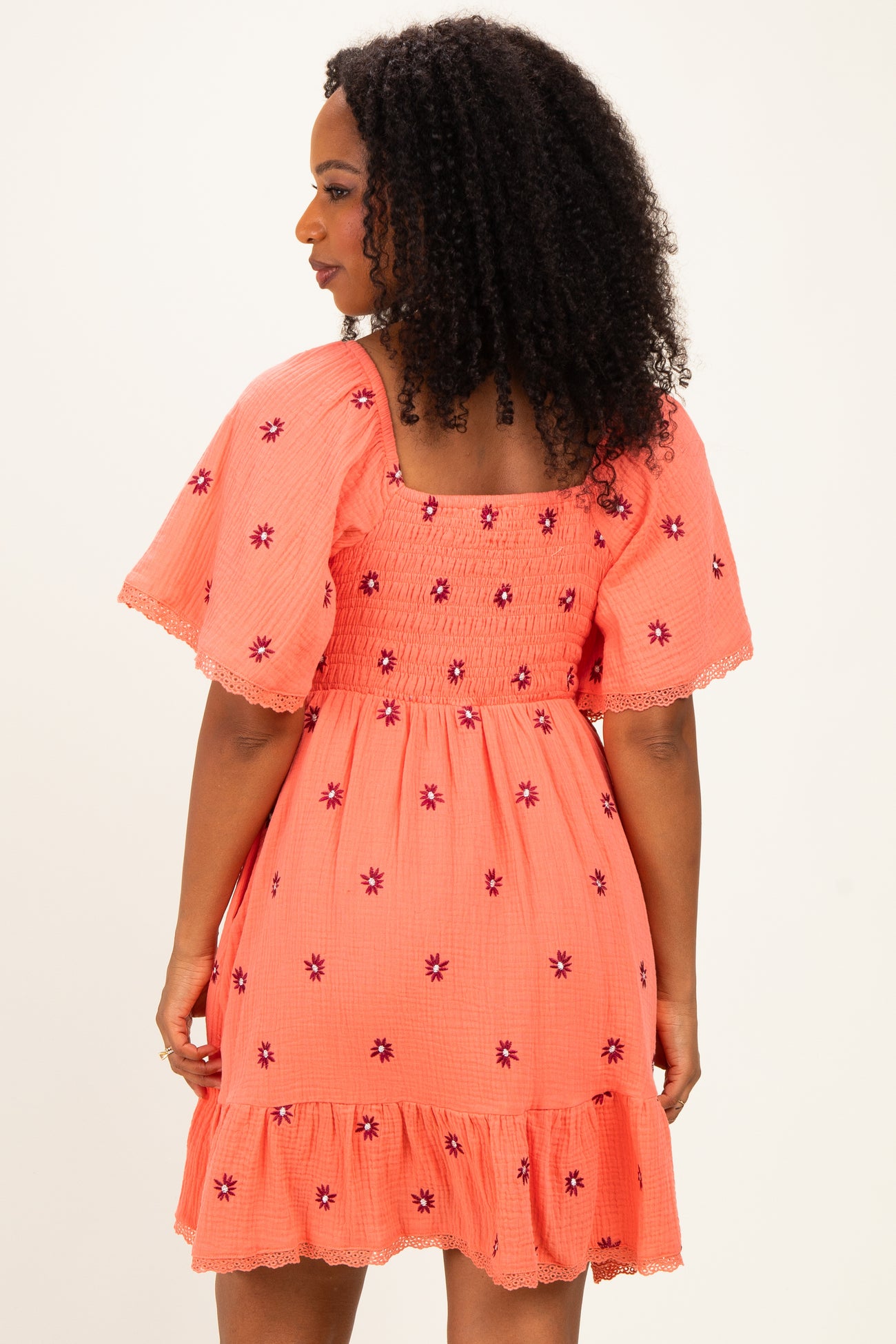 woman wearing a coral dress with floral embroidery and a smocked back, featuring short flutter sleeves and a ruffled hem