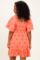 woman wearing a coral dress with floral embroidery and a smocked back, featuring short flutter sleeves and a ruffled hem
