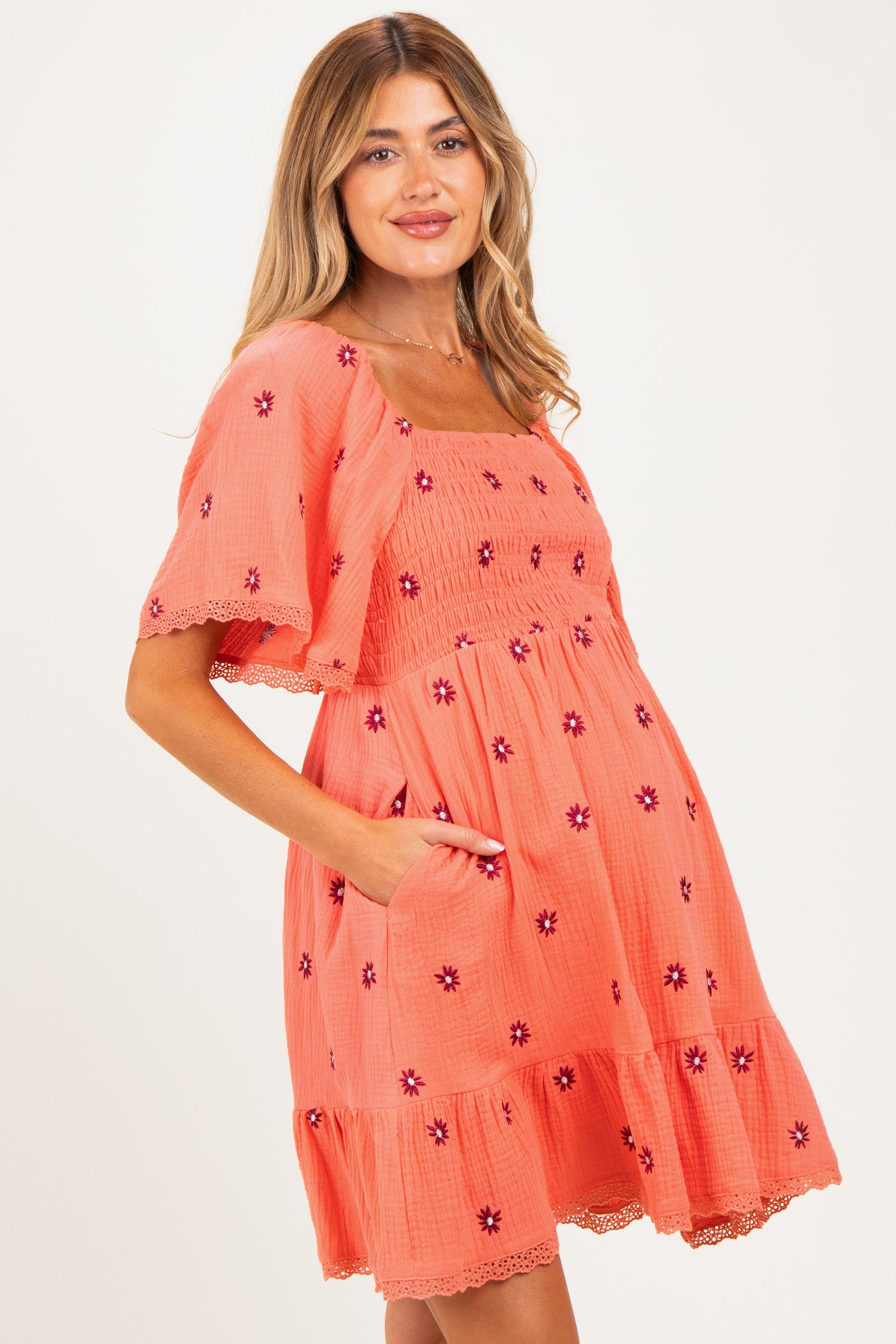 woman wearing a coral maternity dress with short sleeves, floral embroidery, and pockets, showing off her baby bump