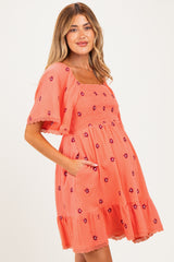 woman wearing a coral maternity dress with short sleeves, floral embroidery, and pockets, showing off her baby bump