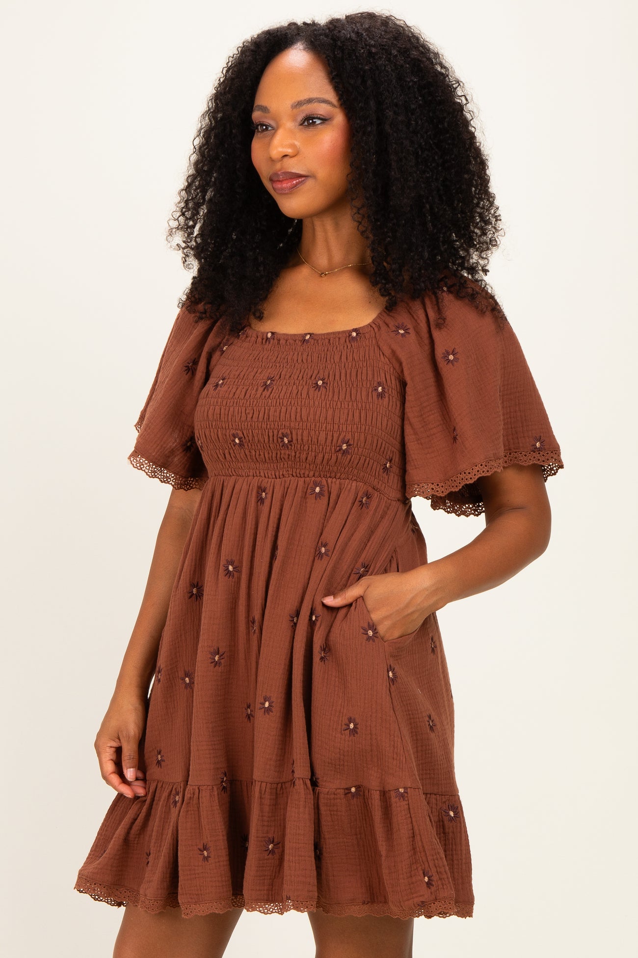 woman wearing a brown short puff sleeve dress with embroidered flowers and a tiered ruffle hem, posing with one hand in pocket