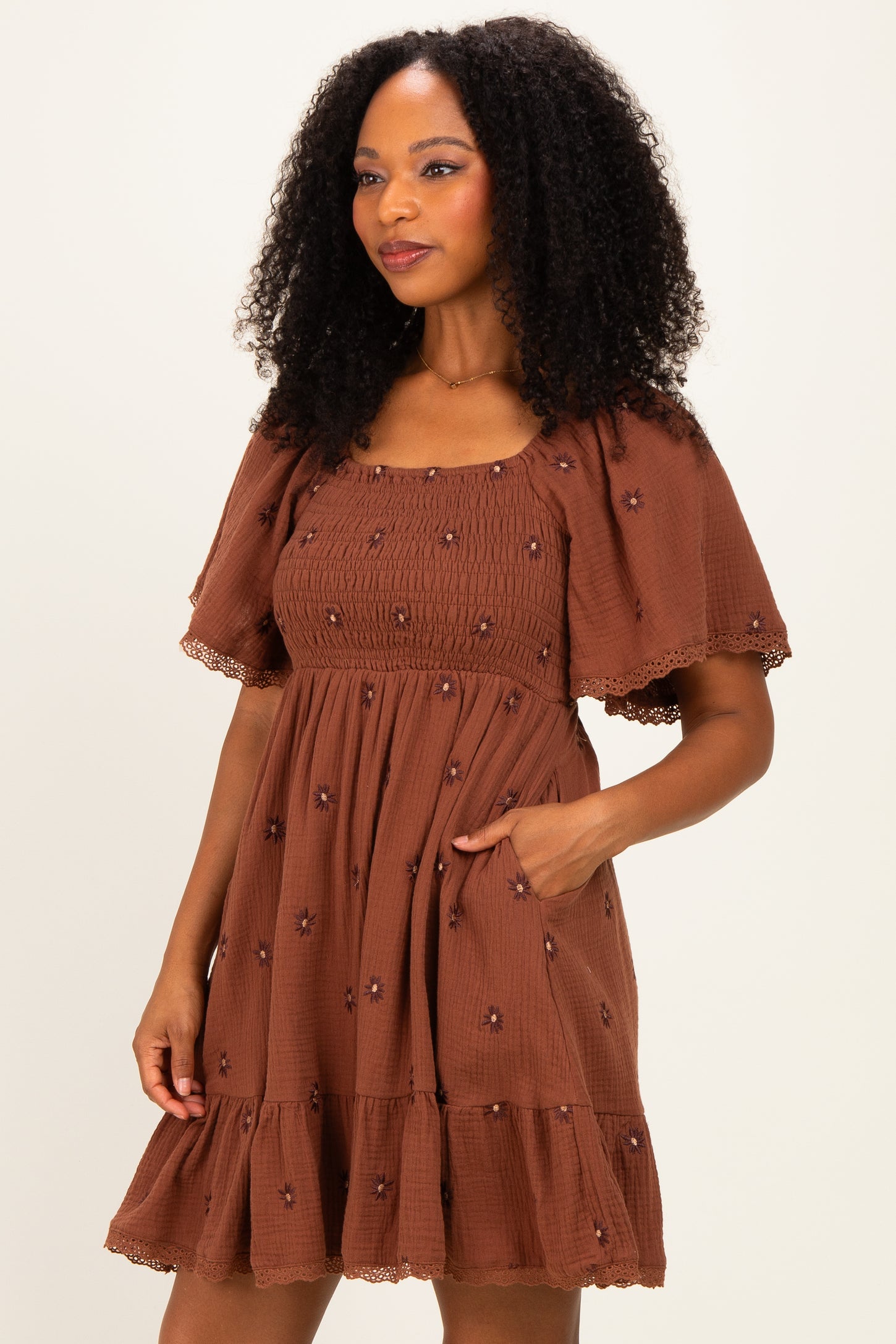 woman wearing a brown short puff sleeve dress with embroidered flowers and a tiered ruffle hem, posing with one hand in pocket