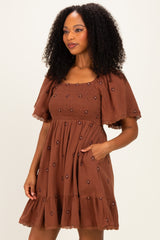 woman wearing a brown short puff sleeve dress with embroidered flowers and a tiered ruffle hem, posing with one hand in pocket