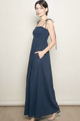 woman wearing a navy blue maxi dress with thin straps and pockets, styled with a smocked bodice