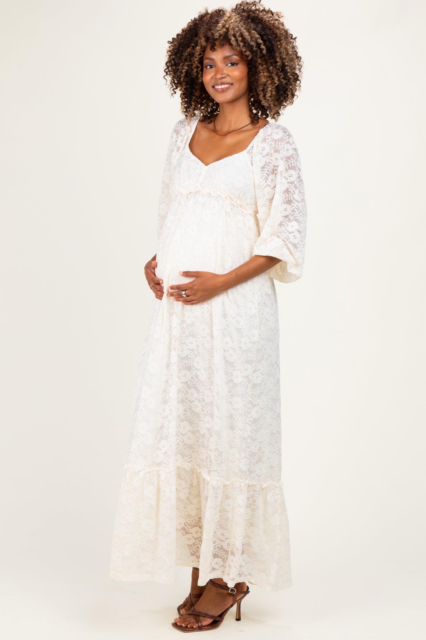 woman wearing a cream lace maternity maxi dress with flutter sleeves and holding her baby bump