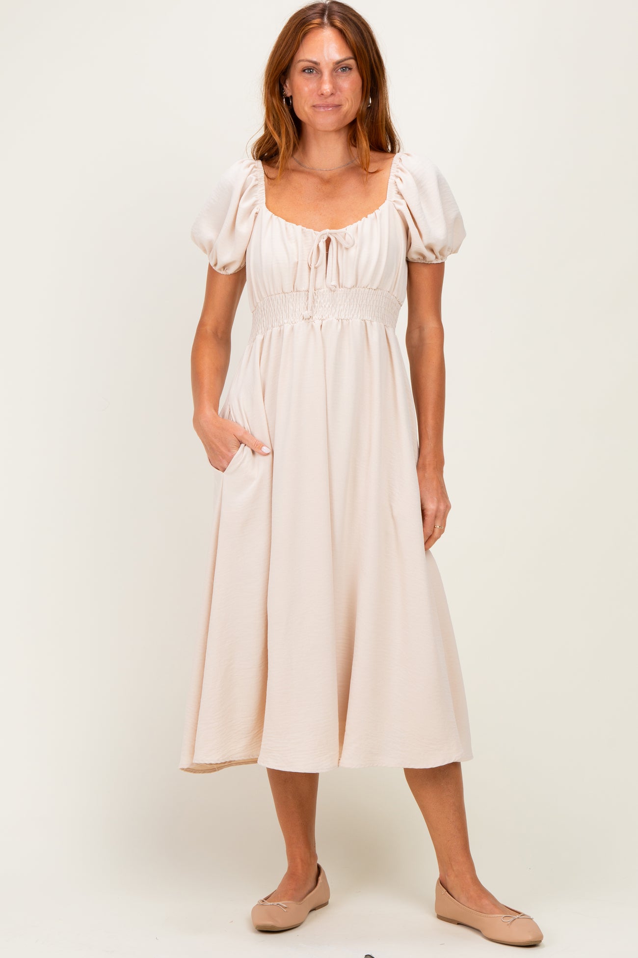 woman wearing a lightweight beige midi dress with puff sleeves and a smocked waist, standing with one hand in the pocket