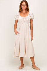 woman wearing a lightweight beige midi dress with puff sleeves and a smocked waist, standing with one hand in the pocket