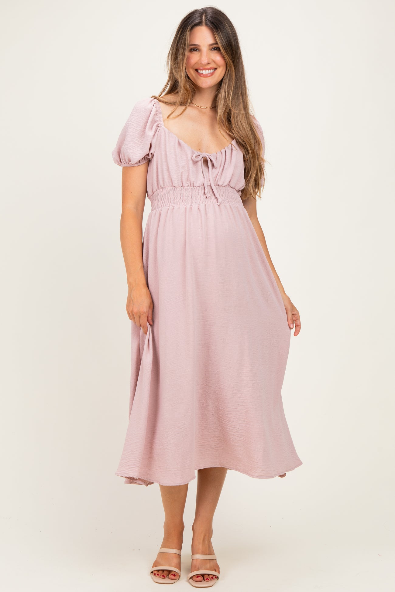 woman wearing a light pink maternity dress with puff sleeves and a tie-front detail, standing with a baby bump visible