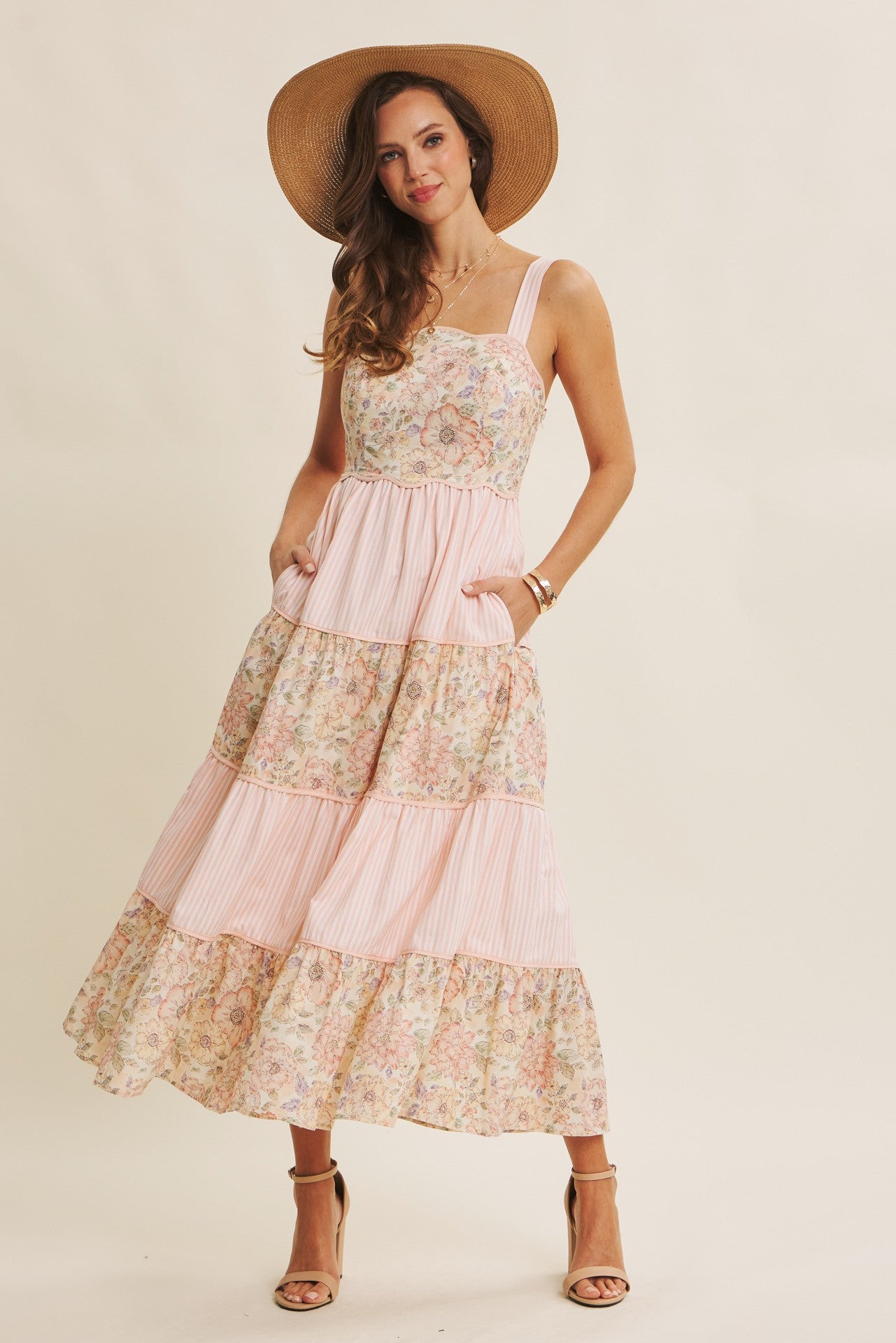 woman wearing a floral print maxi dress with a fitted bodice, tiered skirt, and pockets, paired with a wide-brimmed hat