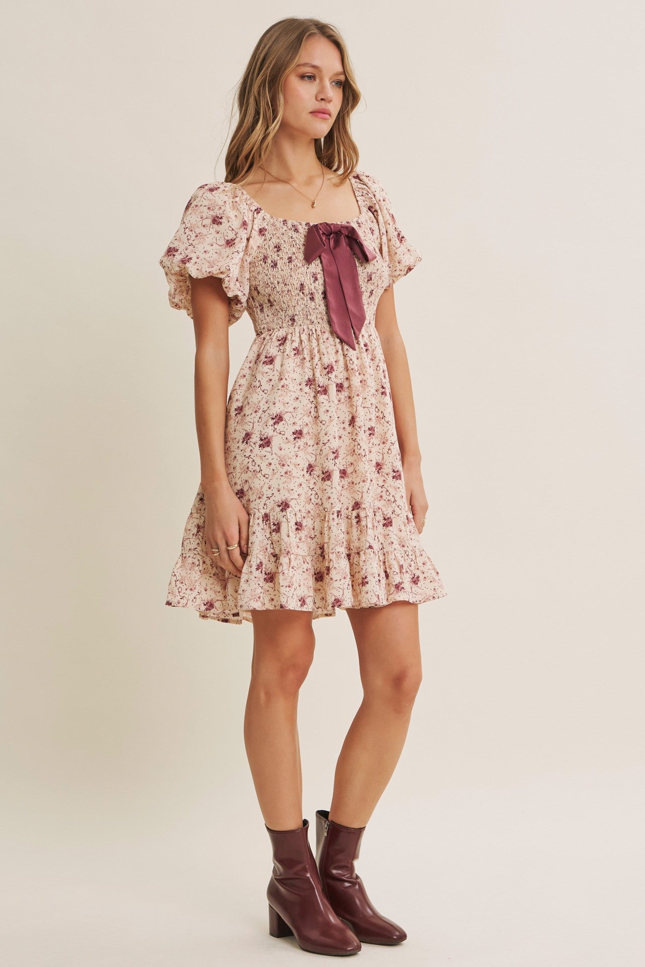 woman wearing a floral print short dress with puff sleeves and a burgundy bow detail, paired with burgundy ankle boots