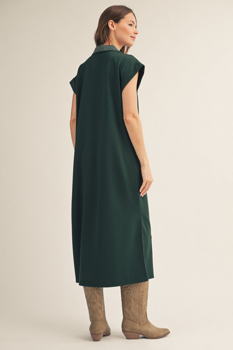 woman wearing a long green sleeveless dress with a collar and knee-high beige boots, standing sideways