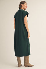 woman wearing a long green sleeveless dress with a collar and knee-high beige boots, standing sideways