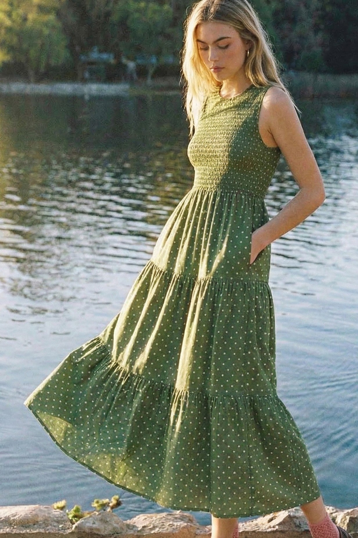 woman wearing a green polka dot maxi dress with a smocked bodice and pockets, standing by a body of water