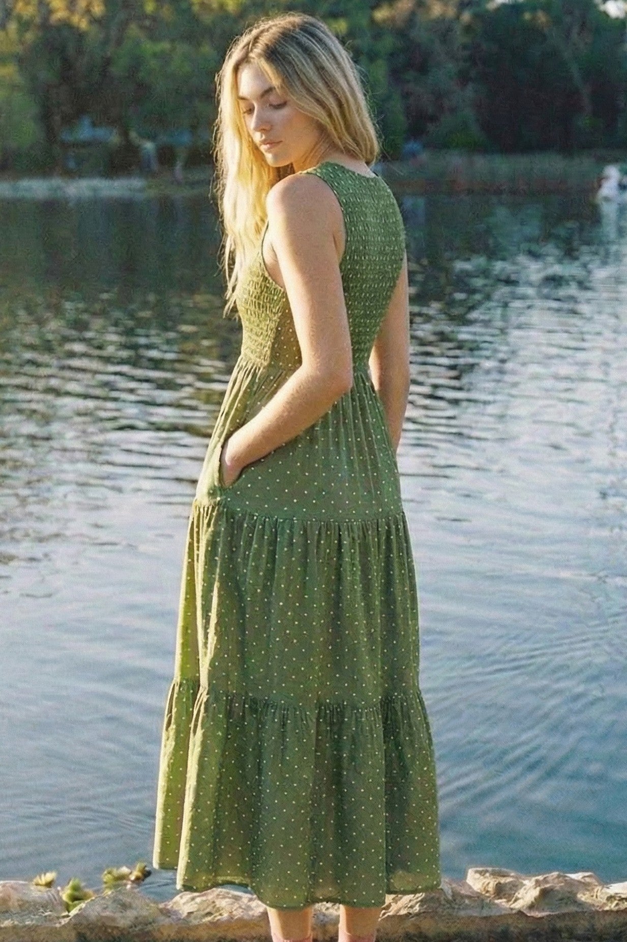 woman wearing a green tiered maxi dress with polka dots and pockets, standing by a lake
