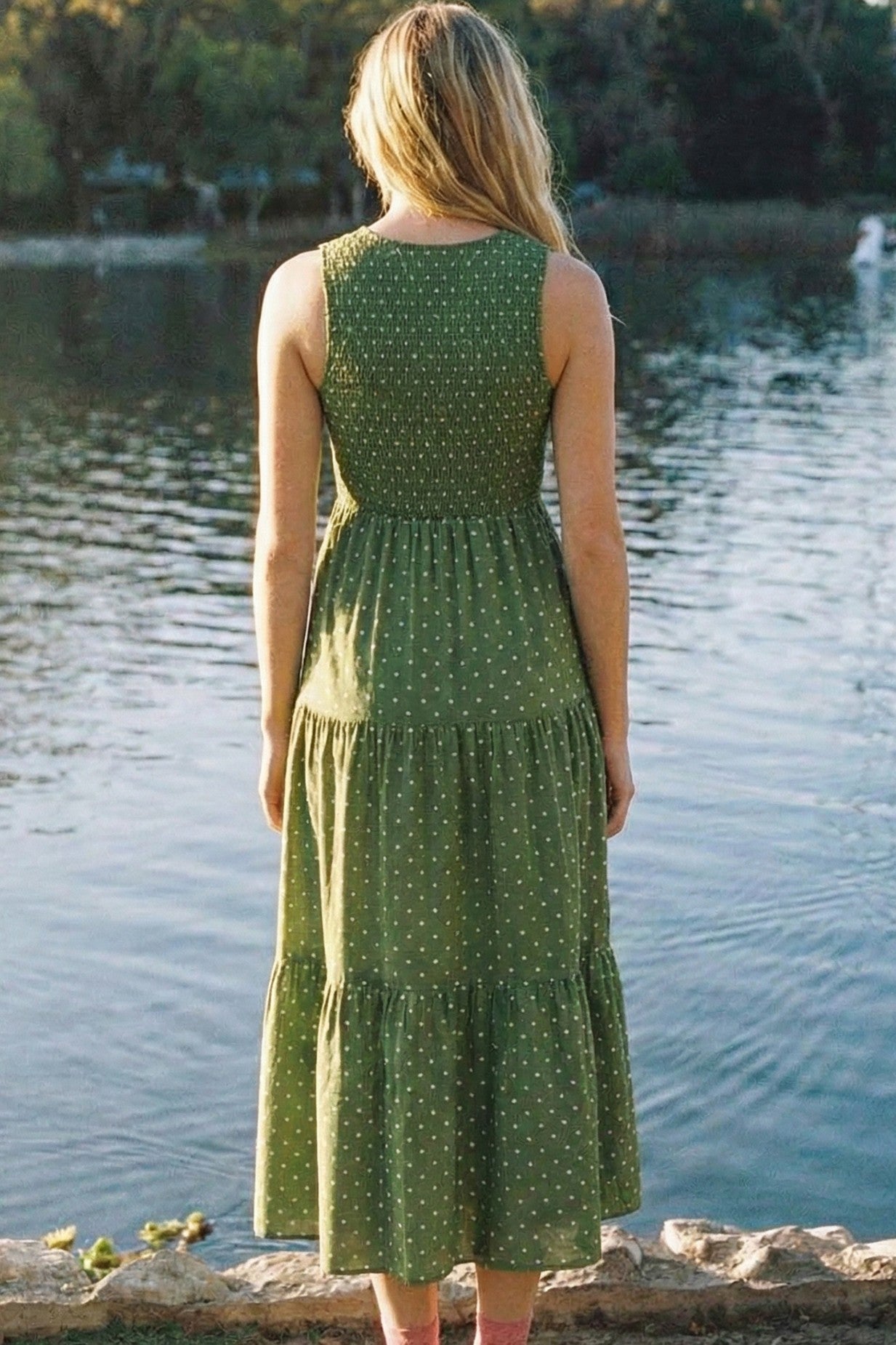 woman wearing a green polka dot tiered midi dress, standing by a lake with her back to the camera
