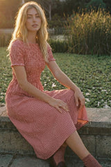woman wearing a red and white gingham print midi dress sitting by a pond with tall grass in the background