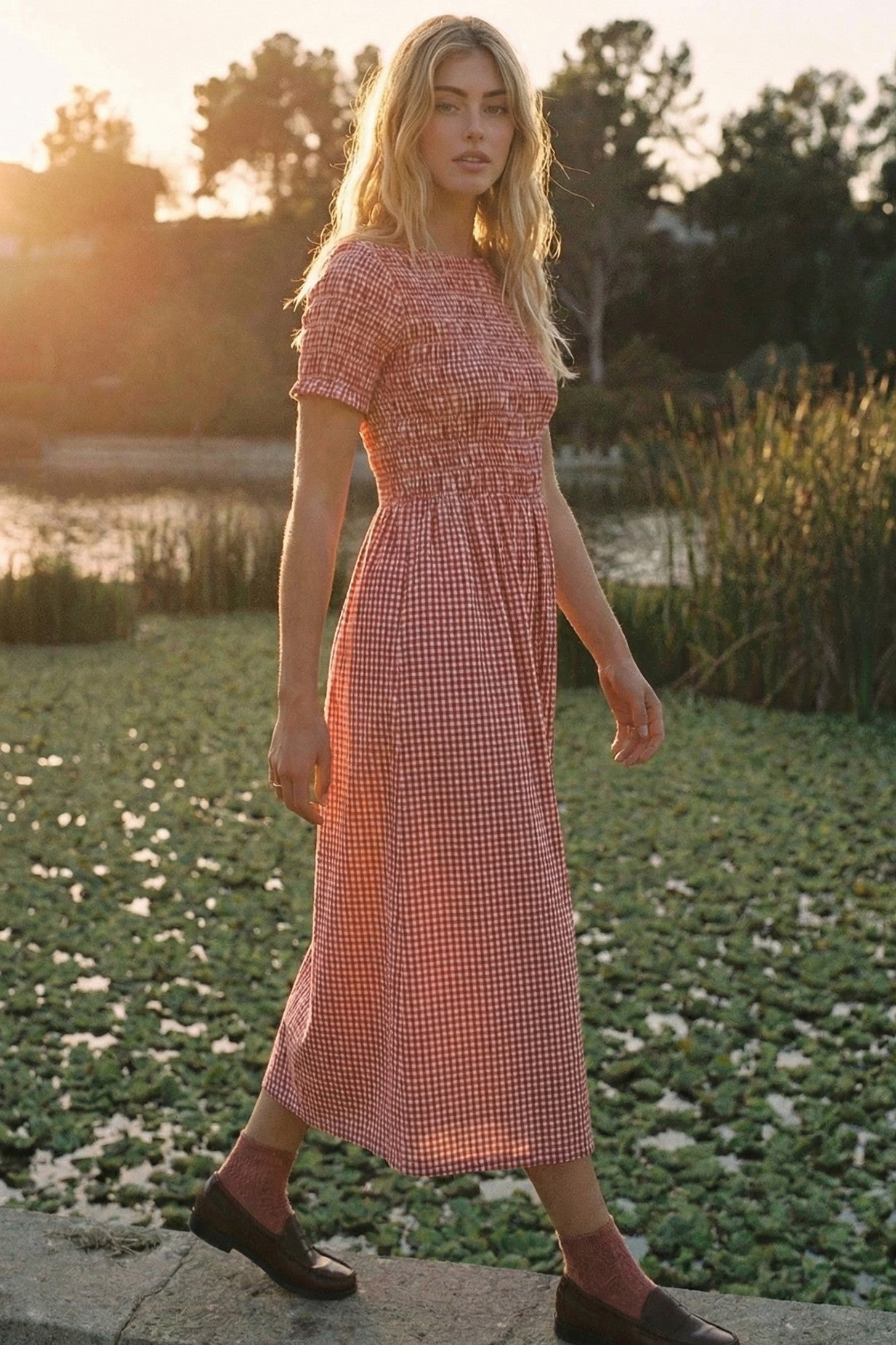 woman wearing a red and white gingham midi dress with short sleeves, standing by a pond during sunset