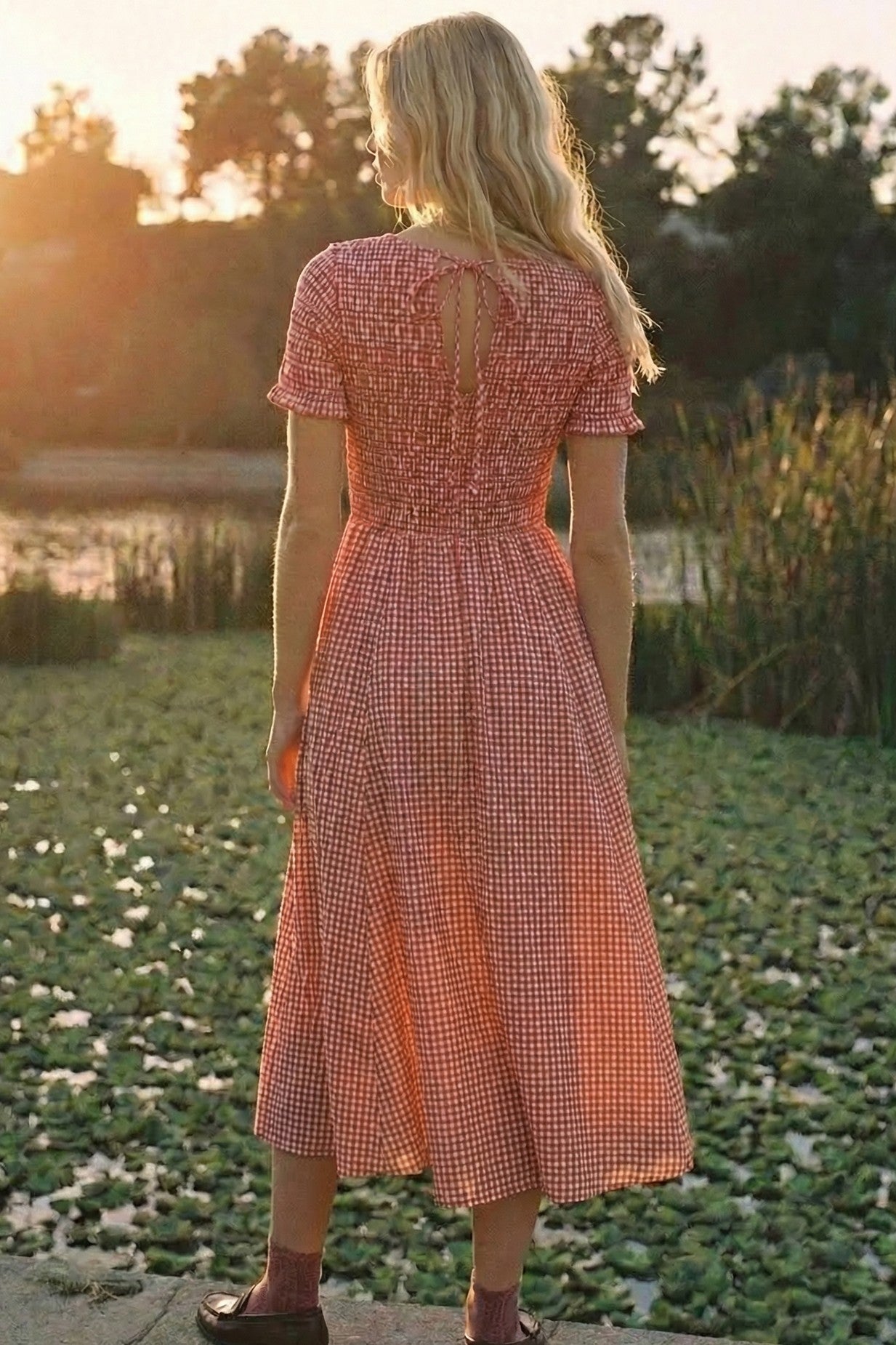 woman wearing a red and white gingham midi dress with a smocked back, standing by a pond at sunset