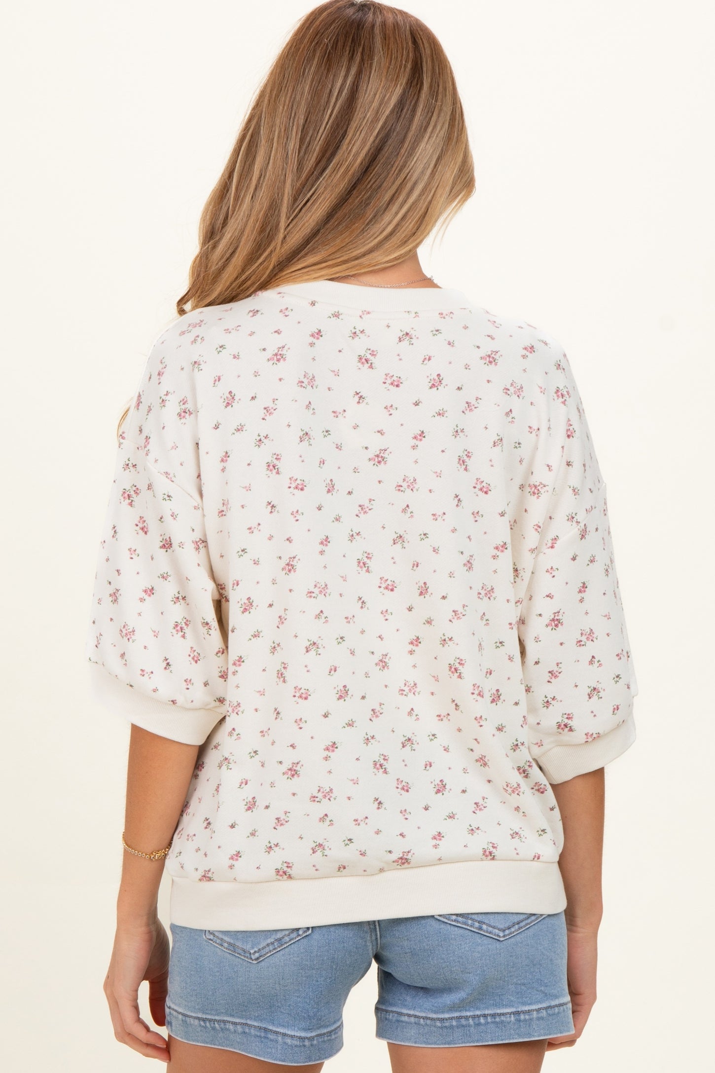 woman wearing a cream floral maternity sweatshirt with three-quarter sleeves, paired with denim shorts