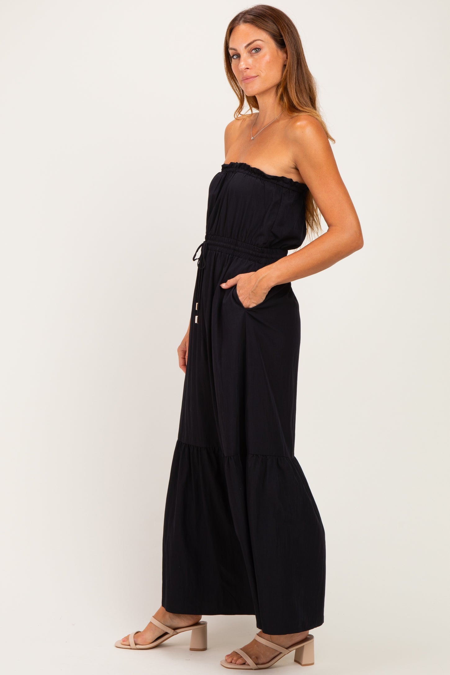 woman wearing a black strapless maxi dress with a tiered skirt and pockets, styled with nude heeled sandals