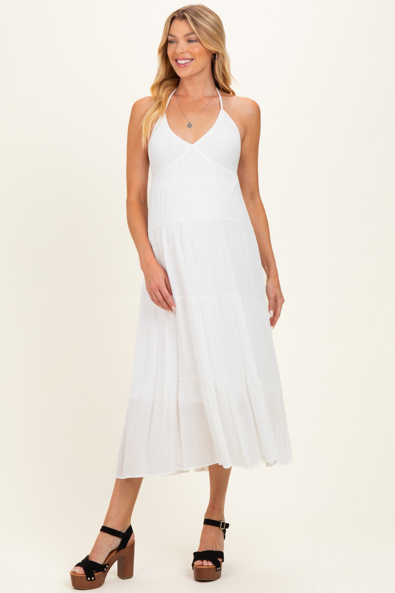 woman wearing a white halter neck maternity dress with tiered skirt and black platform sandals, baby bump not visible