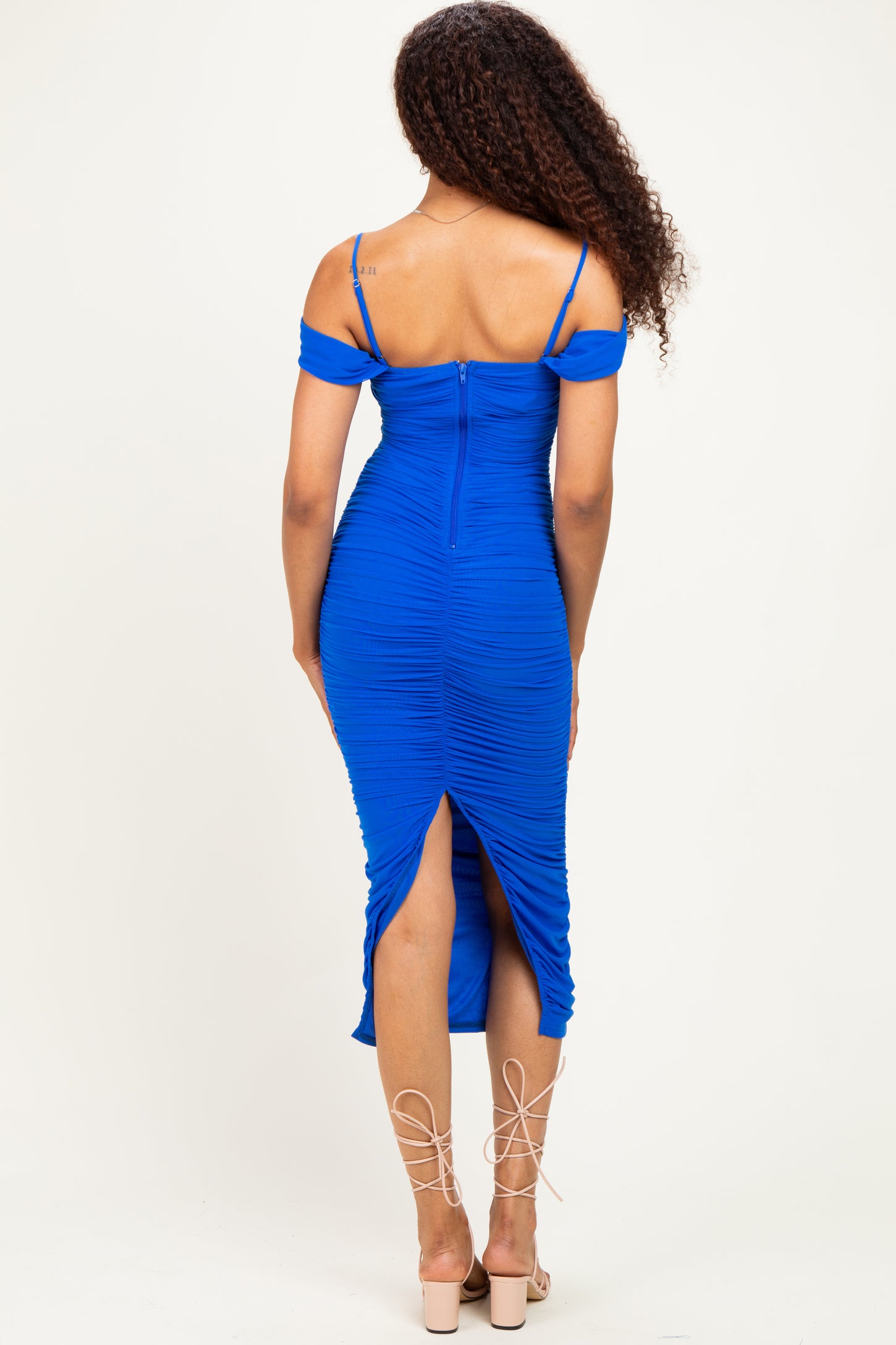 woman wearing a royal blue ruched midi dress with off-the-shoulder sleeves and a side slit, view from the back