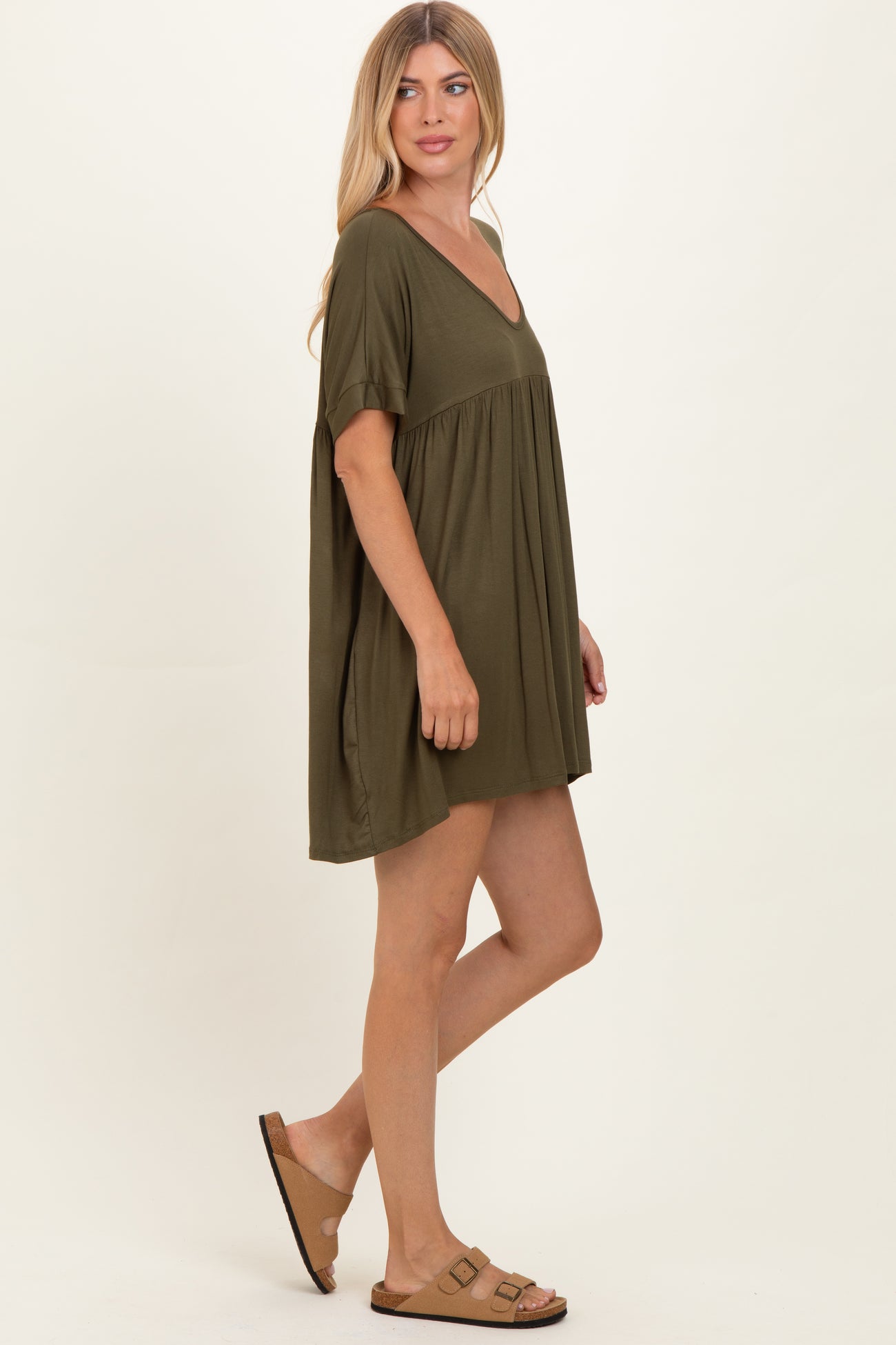 woman wearing an olive green short sleeve swing dress and brown sandals, posing sideways against a neutral background