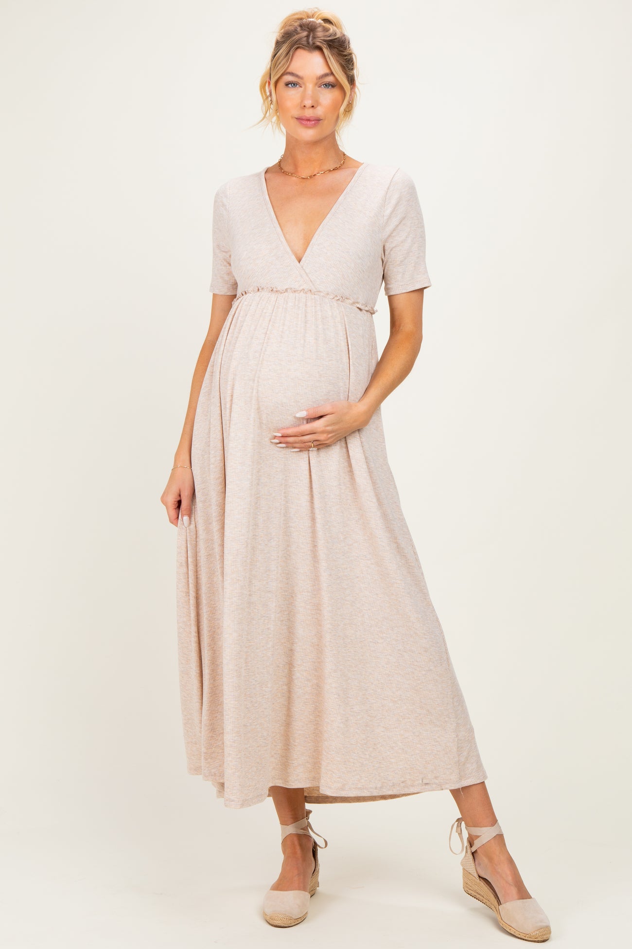 woman wearing a light beige short sleeve maternity dress, holding her baby bump with a flowy skirt and espadrille shoes