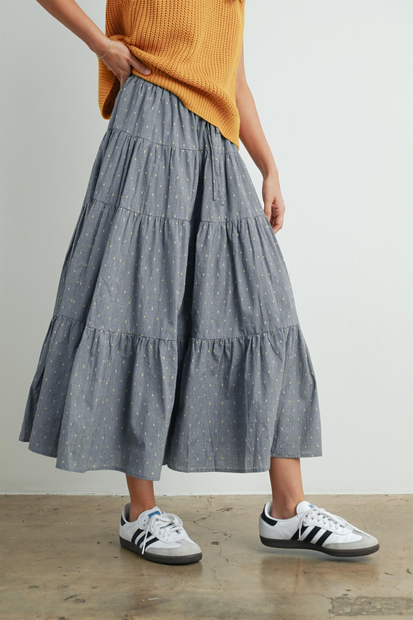 woman wearing a gray tiered midi skirt with yellow polka dots and white sneakers, paired with a relaxed orange sweater
