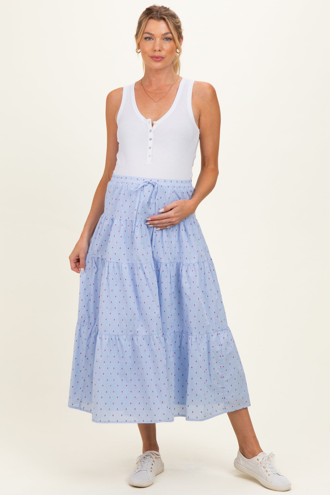 woman wearing a white sleeveless maternity top and a blue tiered maternity skirt with red polka dots, holding her baby bump