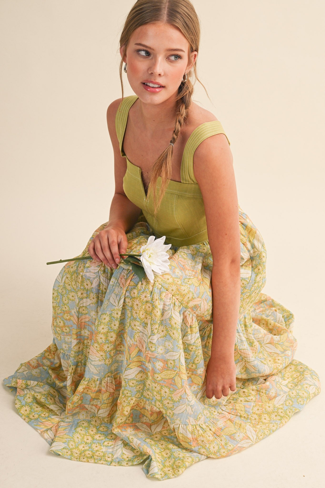 woman wearing a green tank top and a flowy pastel floral maxi skirt, sitting on the floor and holding a white flower