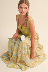 woman wearing a green tank top and a flowy pastel floral maxi skirt, sitting on the floor and holding a white flower