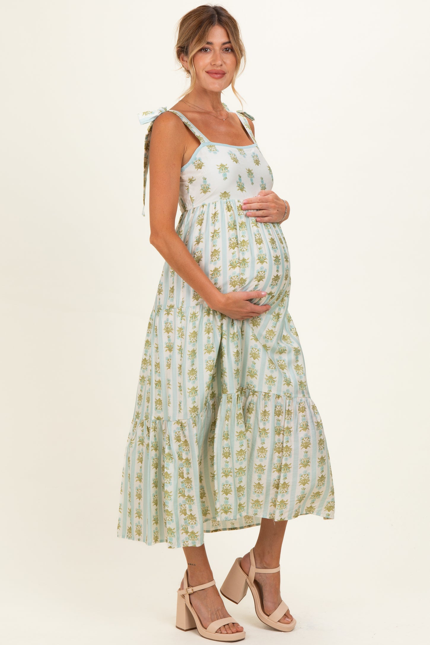 woman wearing a light green floral maternity maxi dress with tie straps, cradling her baby bump