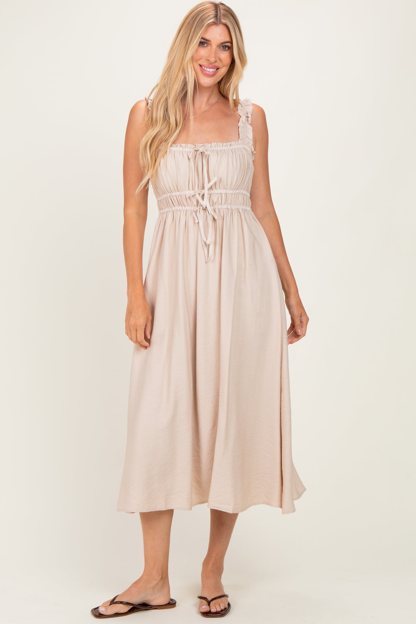 woman wearing a light beige maxi dress with adjustable straps and a drawstring waist, standing and smiling