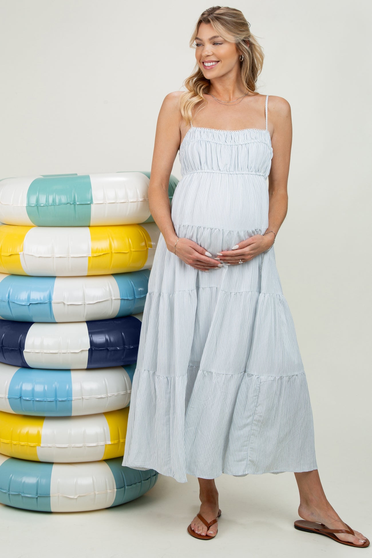 woman wearing a blue and white striped maternity maxi dress with a baby bump, standing beside colorful pool floats