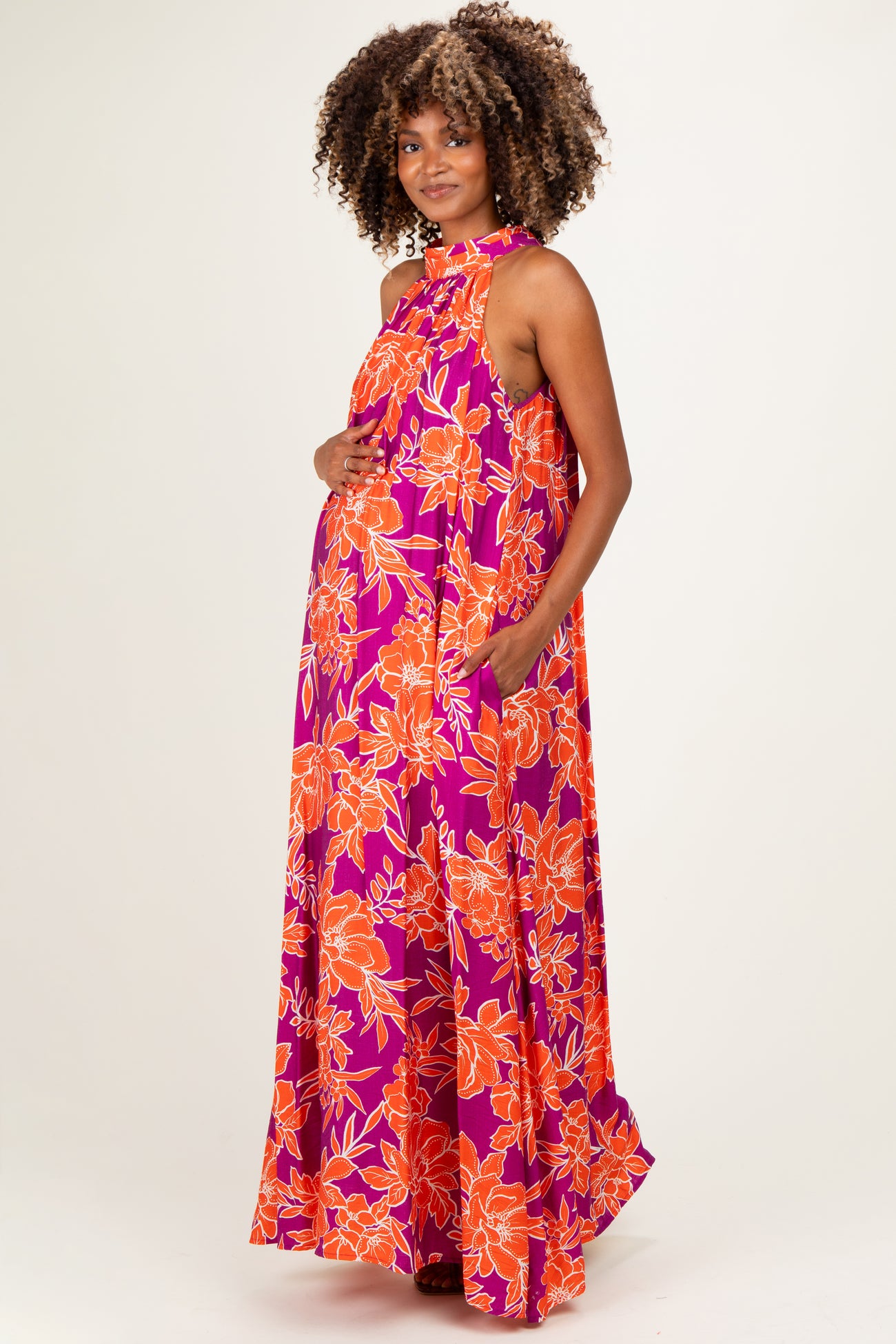 woman wearing a vibrant purple floral maternity maxi dress with a halter neck and pockets, showcasing her baby bump